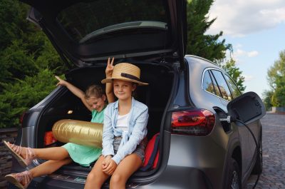Cute little sisters sitting in trunk while waiting for charging car before travelling on summer holiday.